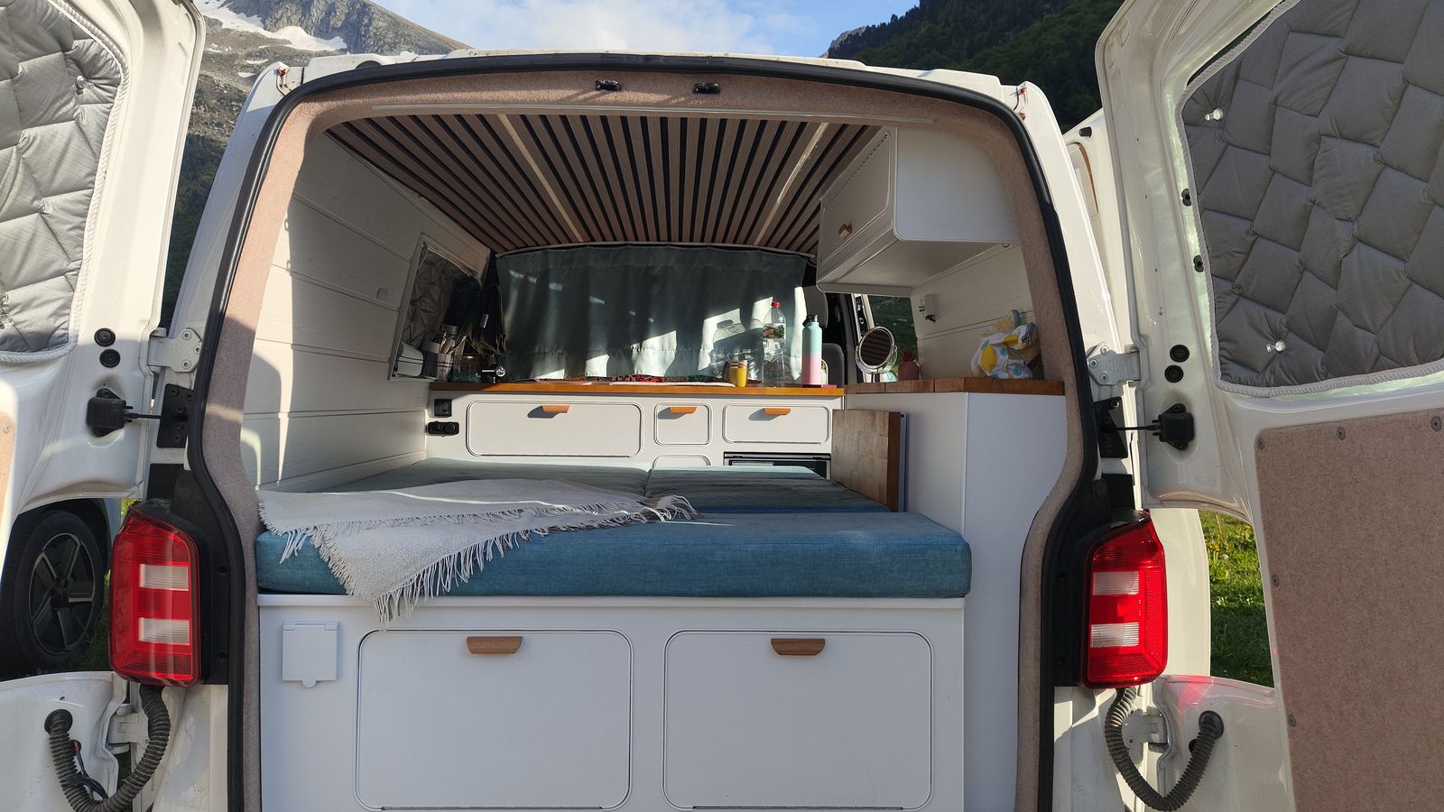 El portón trasero abierto muestra la cama hecha de 180 por 200, los armarios blancos y la encimera de madera al fondo.