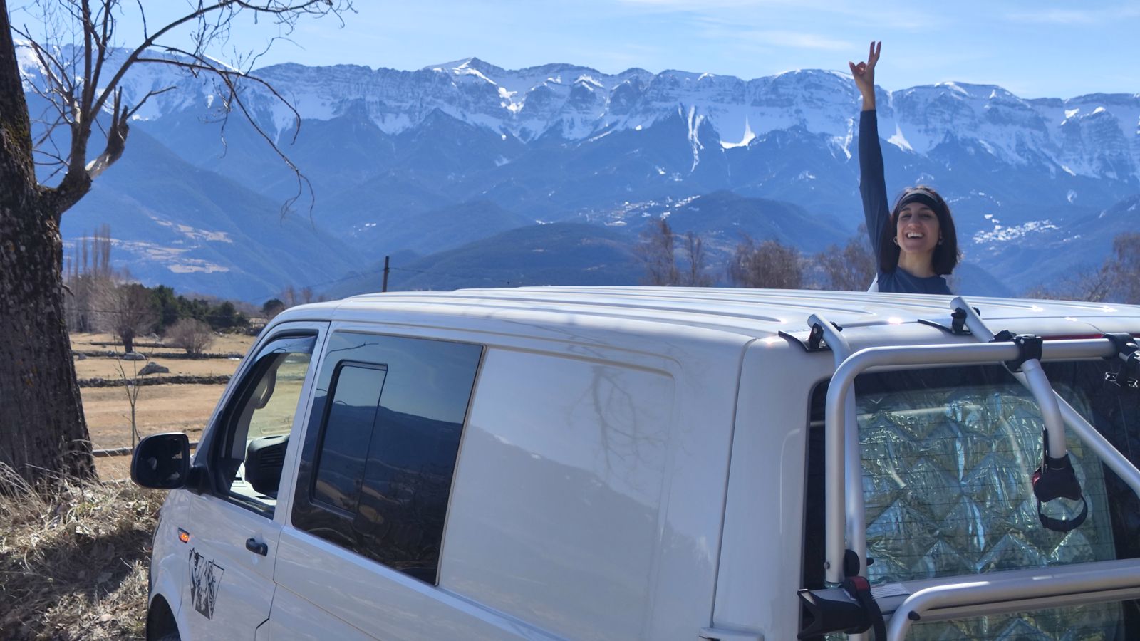 La furgo en un prado del Pirineo con la cordillera nevada al fondo y una viajera celebrando desde el techo.