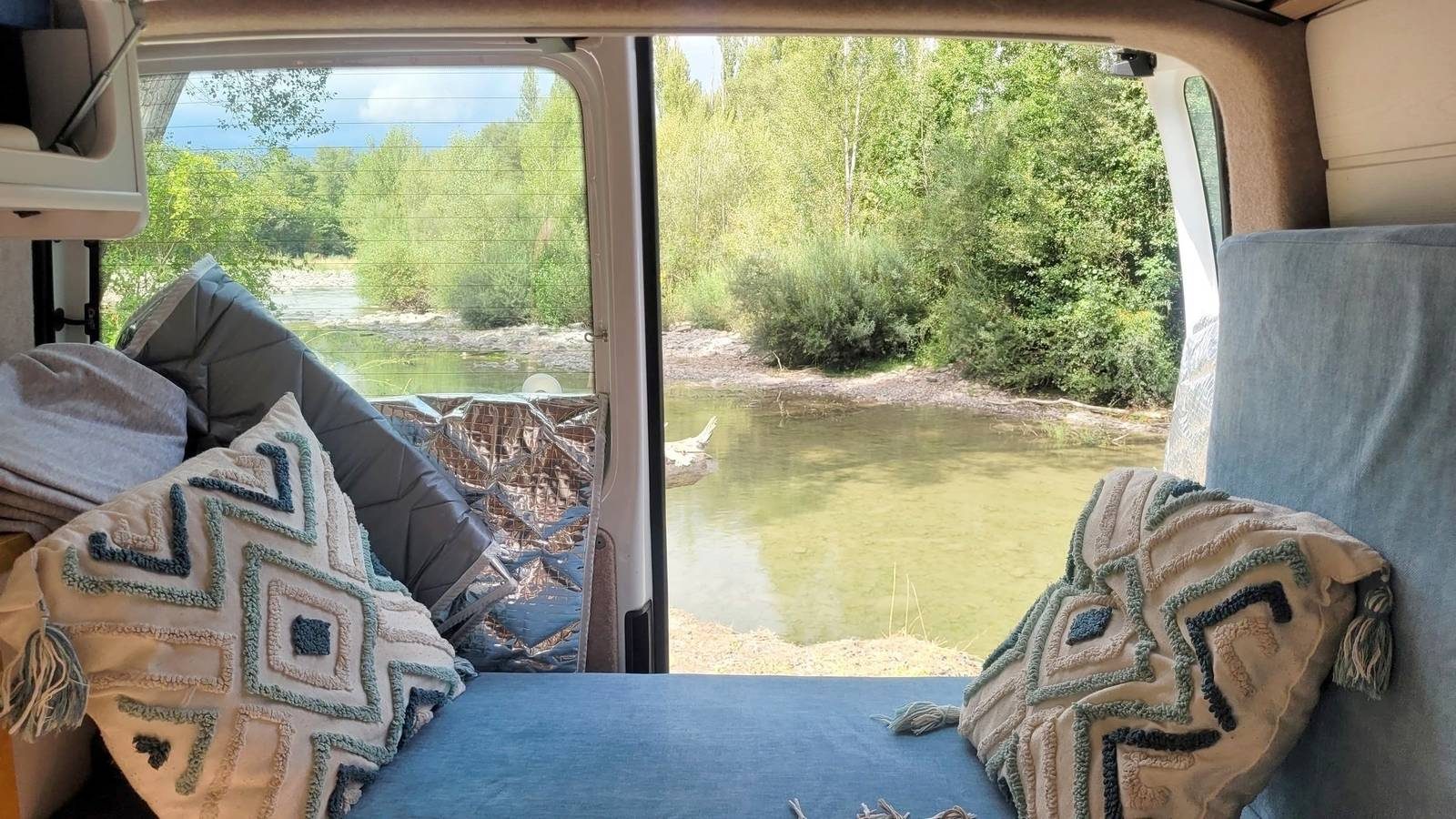 Vista desde la cama de la camper hacia el río, con los cojines y la manta sobre el colchón.